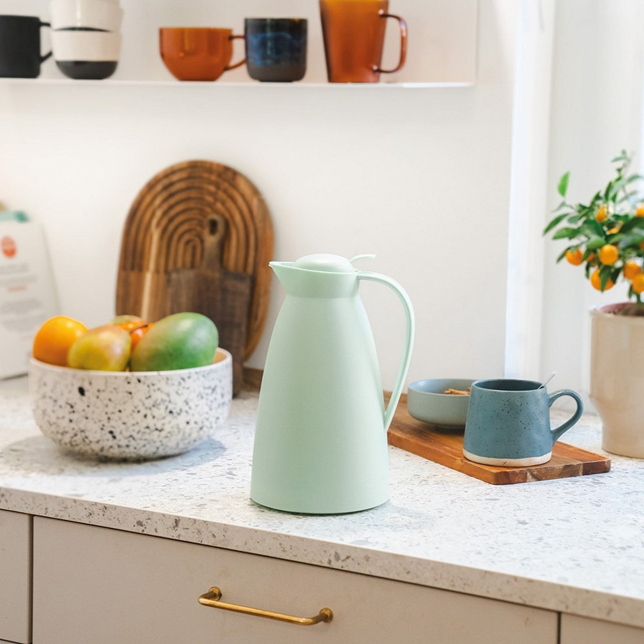 Auf einer Küchenarbeitsplatte steht eine mintgrüne Thermoskanne neben einer Schale mit Obst und einer Tasse. Im Hintergrund sind weitere bunte Tassen auf einem Regal zu sehen, und ein kleines Bäumchen mit orangefarbenen Früchten steht in der Ecke.