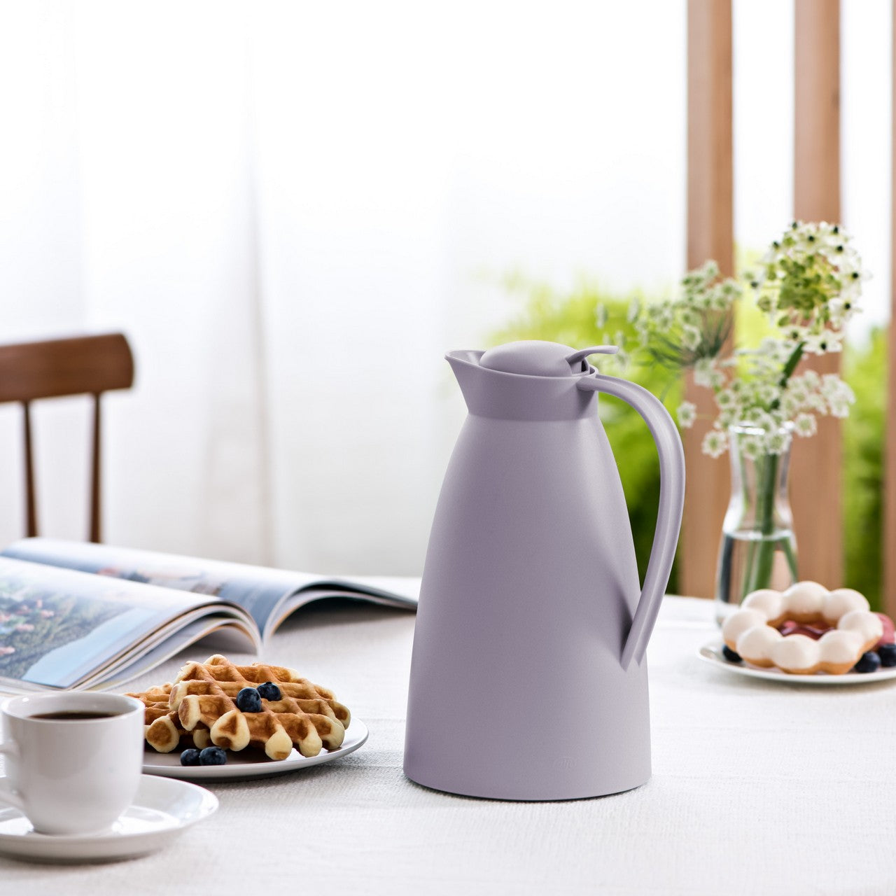 Das Bild zeigt eine lila Thermoskanne und eine Tasse auf einem runden Tisch. Daneben liegt geschnittene Mango auf einem kleinen Brett. Im Hintergrund ist ein Sofa mit einem orangefarbenen Kissen zu sehen, in einem lichtdurchfluteten Raum.