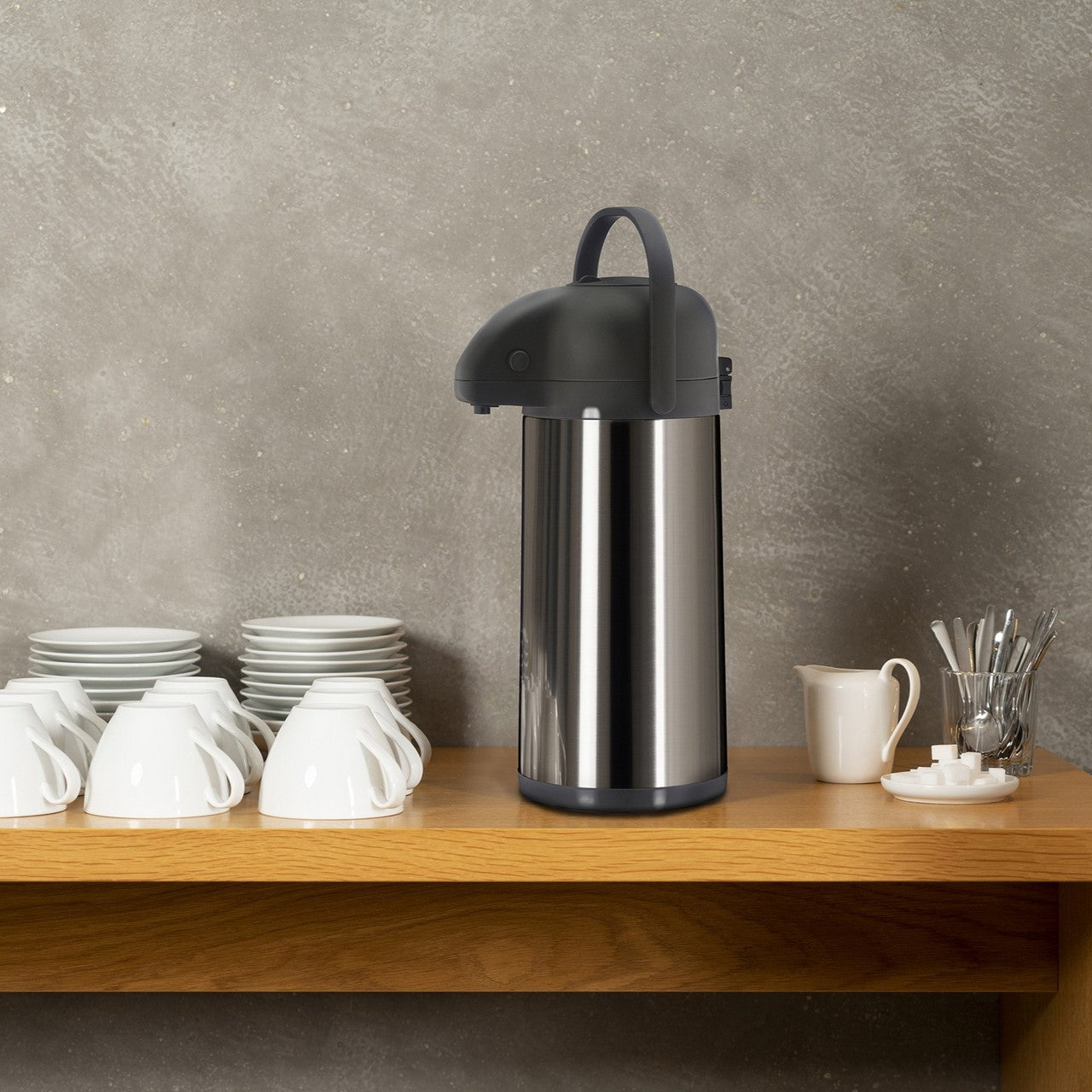 Auf einem Holztisch steht eine große silberne Thermoskanne mit schwarzem Griff. Links sind gestapelte weiße Tassen und Teller, rechts ein kleiner Krug und ein Glas mit Löffeln. Der Hintergrund ist eine graue Betonwand.