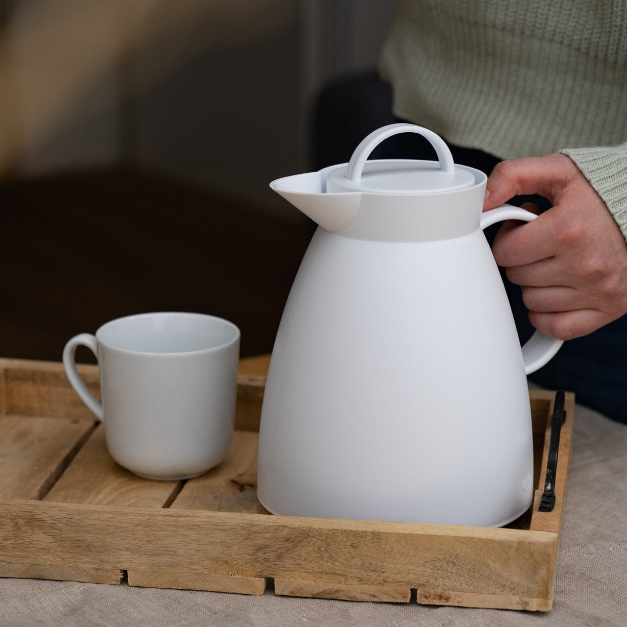 Auf dem Bild ist eine weiße Thermoskanne auf einem Holztablett neben einer weißen Tasse zu sehen. Eine Person mit einem hellgrünen Pullover hält die Kanne am Griff. Der Tisch, auf dem das Tablett steht, hat eine beige Tischdecke.