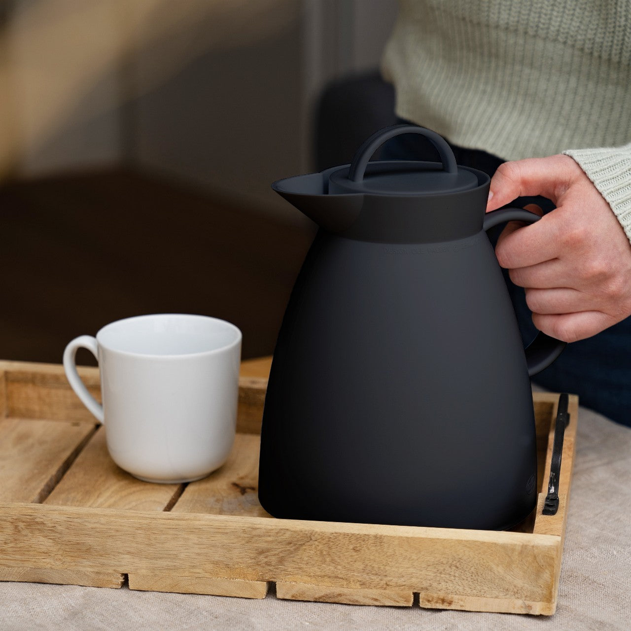 Eine Person hält eine schwarze Kanne auf einem Holztablett, das auch eine weiße Tasse enthält. Das Tablett steht auf einem Tisch, und die Person trägt einen hellen Strickpullover.