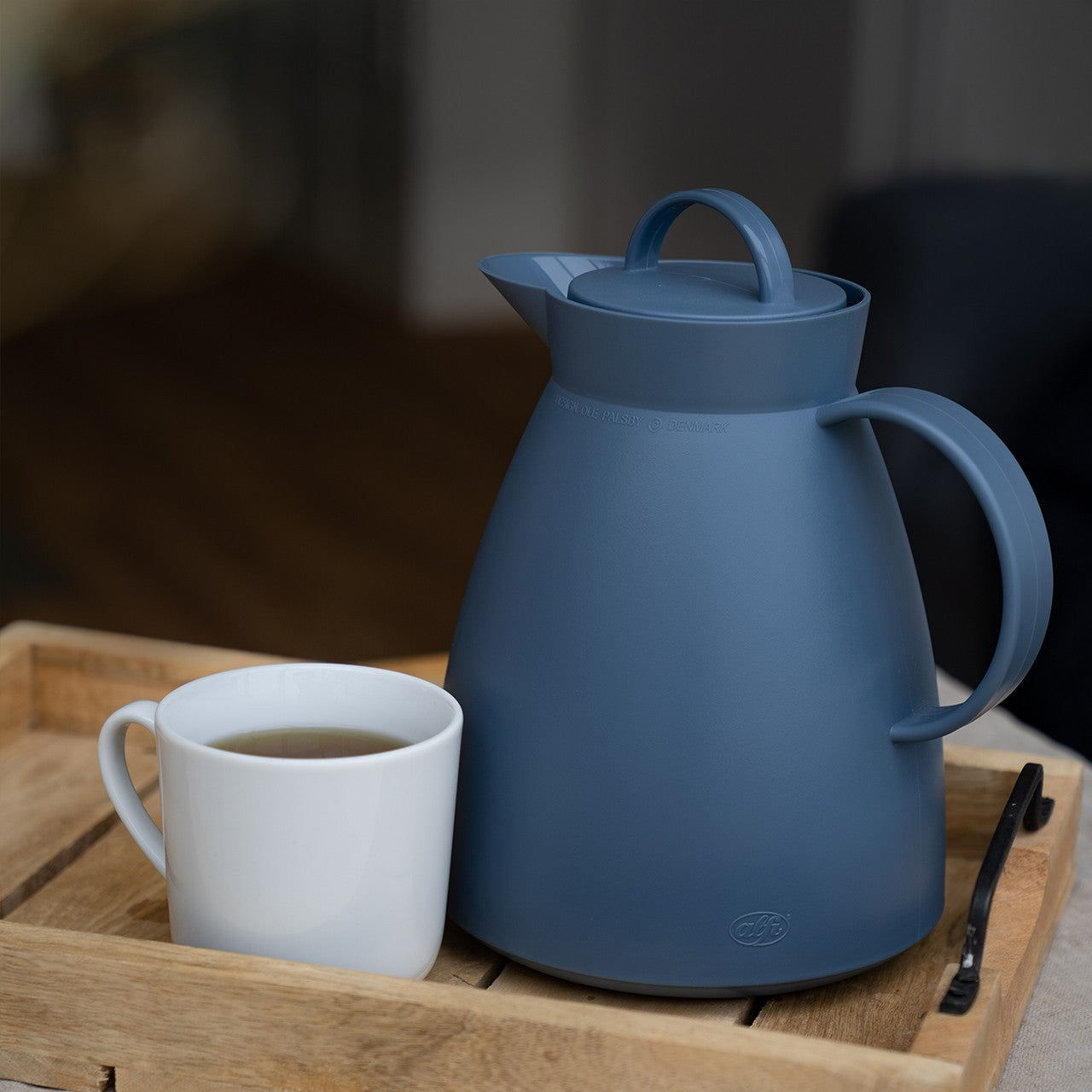 Eine blaue Thermoskanne steht auf einem Tablett neben einer weißen Tasse mit Heißgetränk. Die Szene wirkt gemütlich und einladend. Im Hintergrund ist ein unscharfes, wohnliches Umfeld.