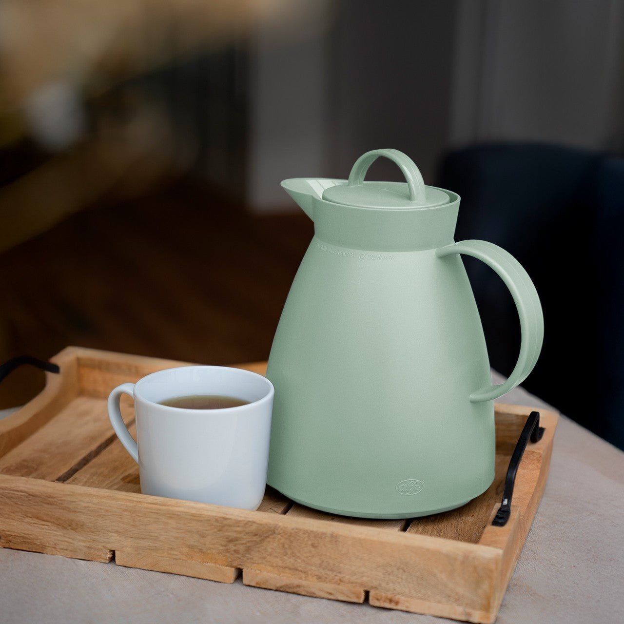 Auf dem Bild befindet sich eine helle, mintgrüne Thermoskanne auf einem Holztablett. Daneben steht eine weiße Tasse mit einem heißen Getränk. Die Szenerie wirkt gemütlich und einladend.