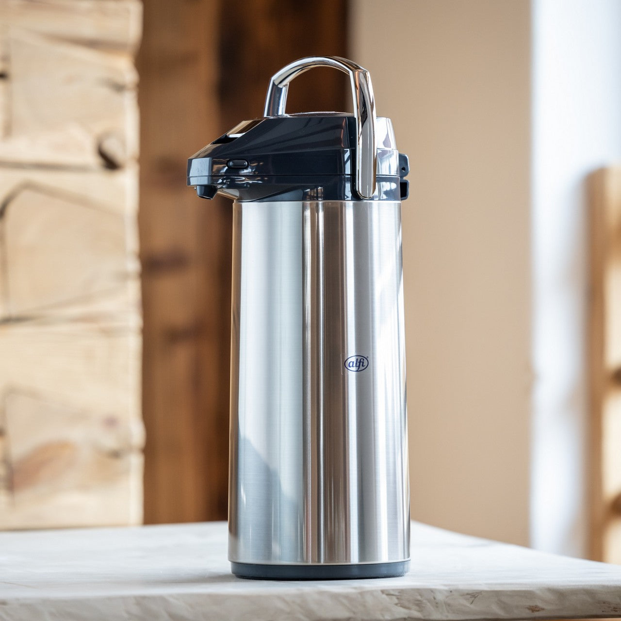 Das Bild zeigt einen silbernen Thermosbehälter mit Pumpmechanismus und einem schwarzen Deckel. Er steht auf einem Tisch vor einer Holzwand. Die Umgebung wirkt gemütlich und rustikal.