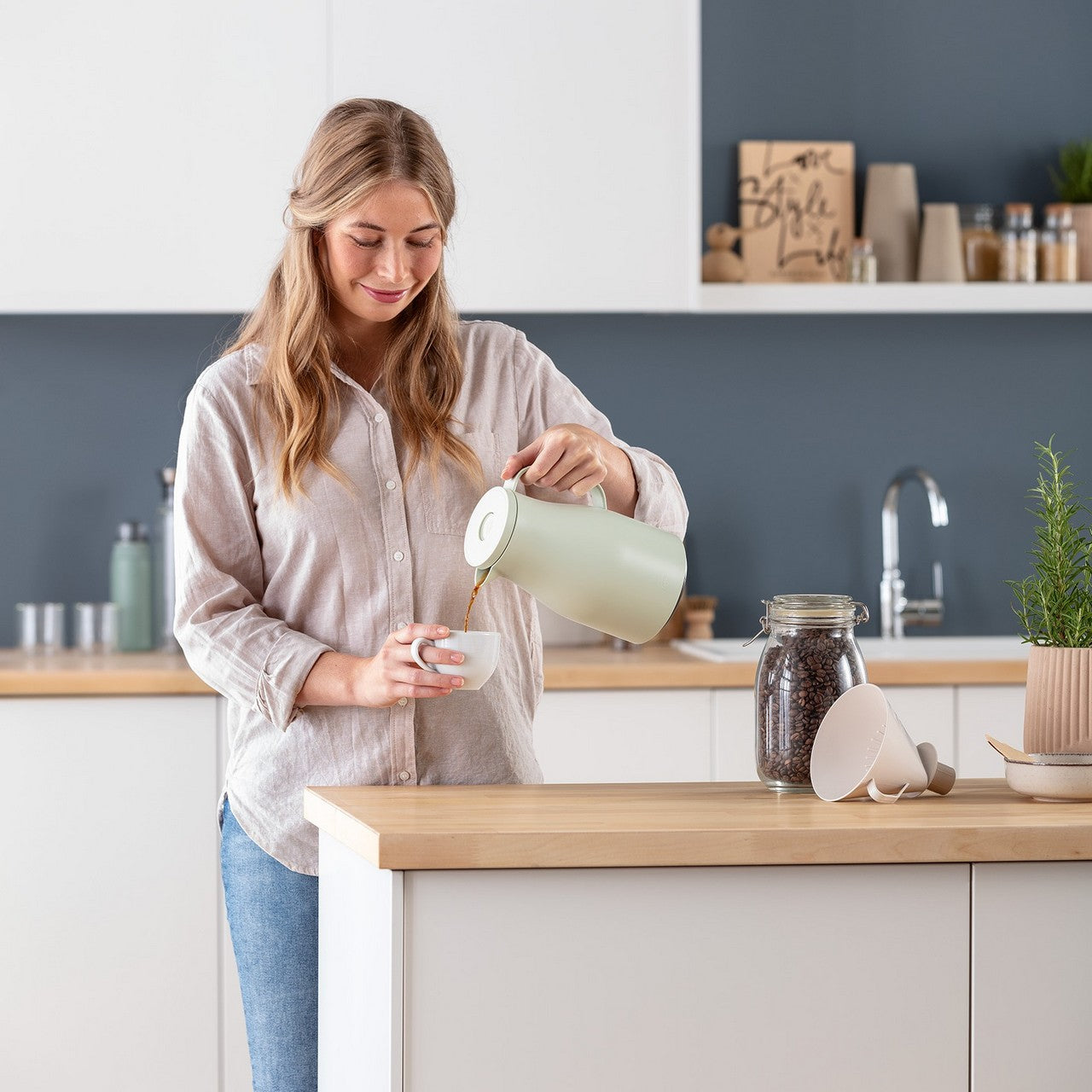 Eine Frau gießt Kaffee aus einem pastellgrünen Kessel in eine Tasse. Auf der Arbeitsplatte stehen ein Glas mit Kaffeebohnen und Filter. Im Hintergrund sind eine Spüle und Regale.