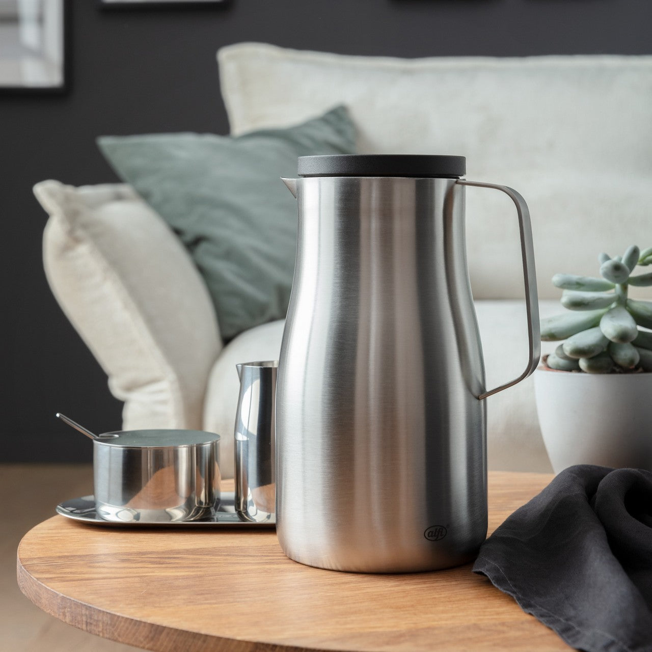 Auf dem Bild steht eine silberne Thermoskanne auf einem Holztisch. Daneben sind ein Milchkännchen und eine Zuckerdose auf einem Tablett sowie ein schwarzes Tuch. Im Hintergrund ist ein Sofa mit grünen Kissen.