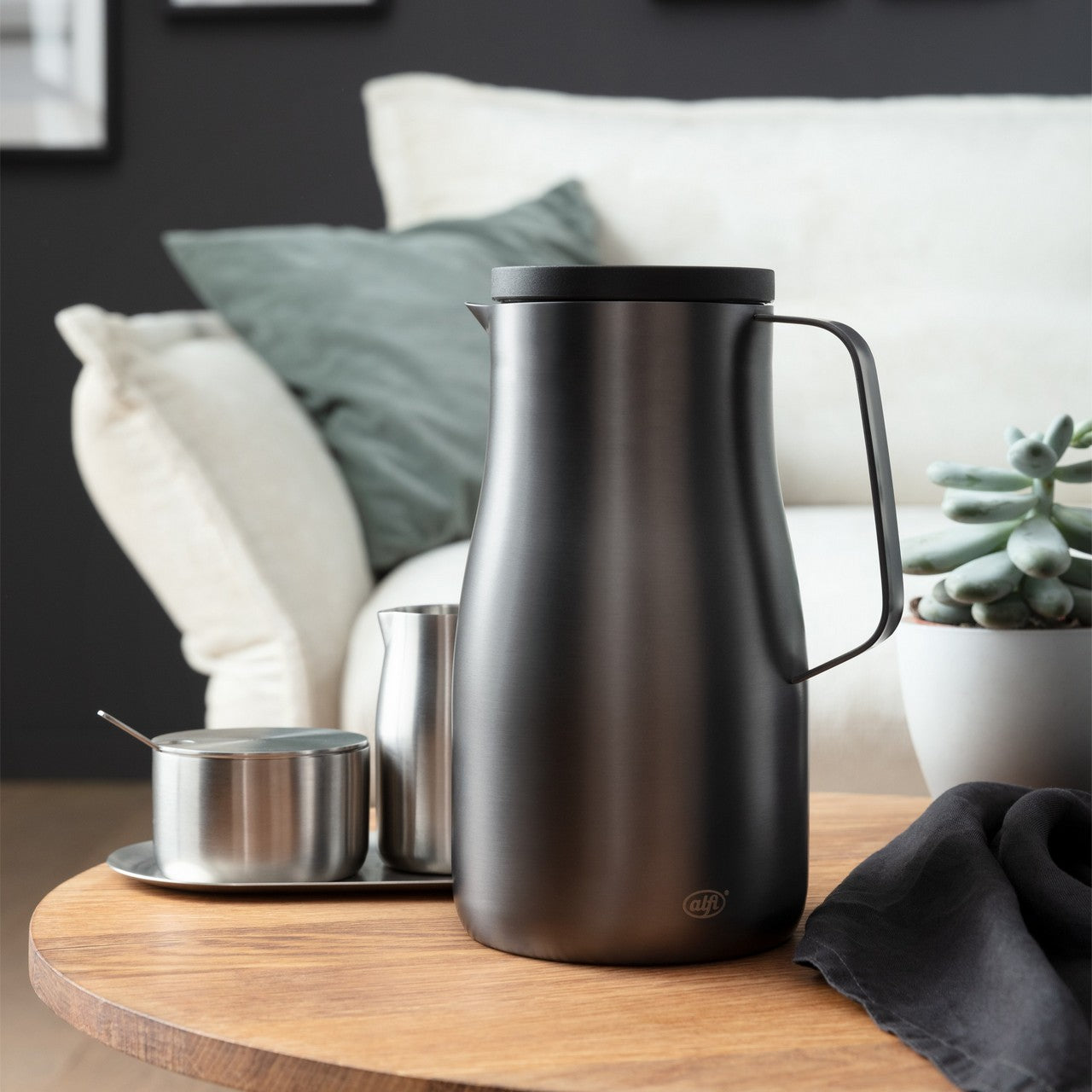 Auf einem Holztisch steht eine matte Edelstahlkanne mit Griff im Vordergrund. Daneben sind ein kleinerer Edelstahlbehälter mit Löffel und ein Becher auf einem Tablett. Im Hintergrund sind ein helles Sofa mit Kissen und eine Pflanze zu sehen.
