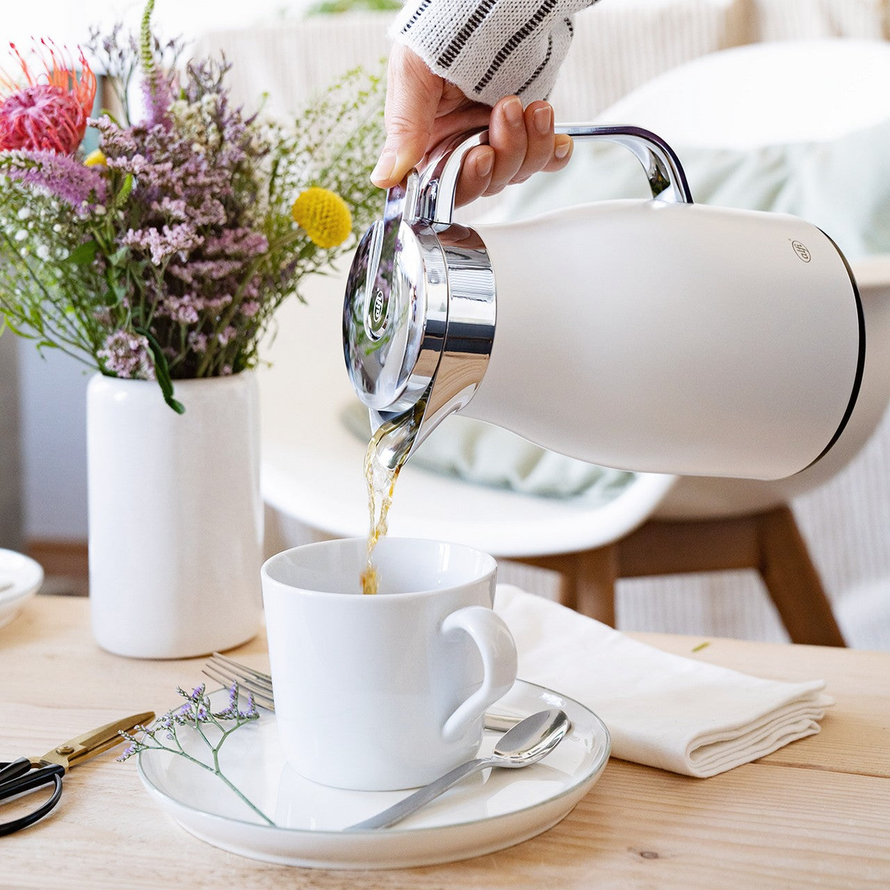 Jemand gießt aus einer eleganten, weißen Teekanne Tee in eine weiße Tasse, die auf einem Tisch steht. Im Hintergrund steht eine Vase mit bunten Blumen. Daneben sind eine Serviette und ein Teelöffel auf dem Tisch platziert.