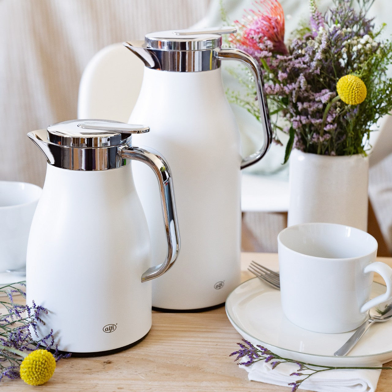 Auf einem Tisch stehen zwei weiße Isolierkannen mit glänzenden Griffen und Deckeln. Daneben sind eine weiße Tasse und ein Teller mit Besteck zu sehen. Im Hintergrund befinden sich getrocknete Blumen in einer Vase.