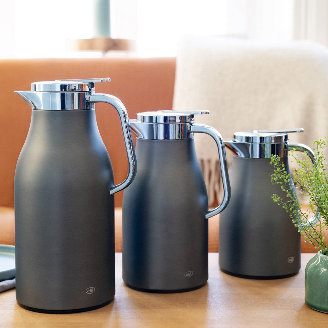 Das Bild zeigt drei moderne, dunkelgraue Thermoskannen mit silbernen Deckeln und Griffen nebeneinander auf einem Tisch. Im Hintergrund sind ein Sofa mit Decke und eine Pflanze zu sehen.