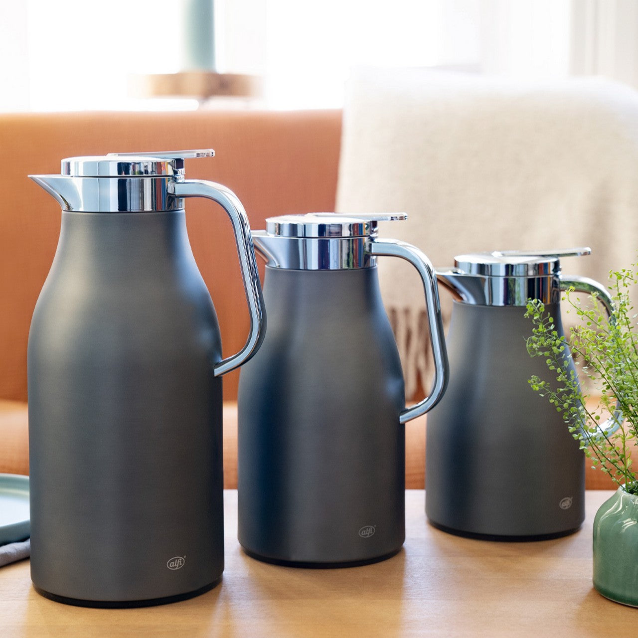 Auf dem Bild sind drei Thermoskannen in verschiedenen Größen auf einem Tisch zu sehen. Die Kannen sind dunkel mit silbernen Deckeln und Henkeln. Im Hintergrund sind unscharf ein Sofa und ein grüner Pflanzenzweig.