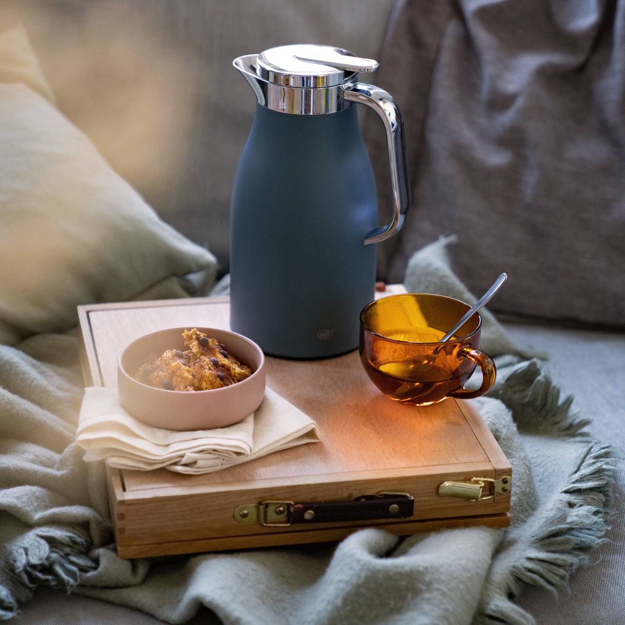 Eine dunkelblaue Thermoskanne und eine bernsteinfarbene Tasse stehen auf einem Holzkasten. Daneben liegt eine Schüssel mit Gebäck auf einem Tuch. Die Szenerie wirkt gemütlich und einladend.