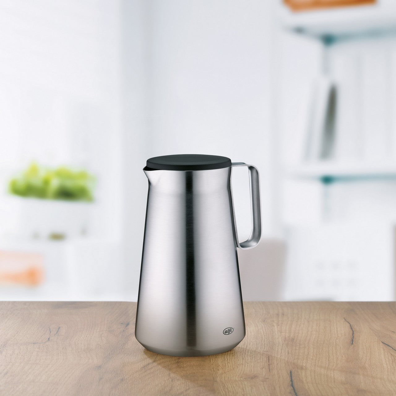 Das Bild zeigt einen glänzenden, silbernen Wasserkocher mit schwarzem Griff auf einer Holzoberfläche in einer modernen Küche. Im Hintergrund sind ein kleines Regal mit grünen Äpfeln und weiße Wände zu sehen.