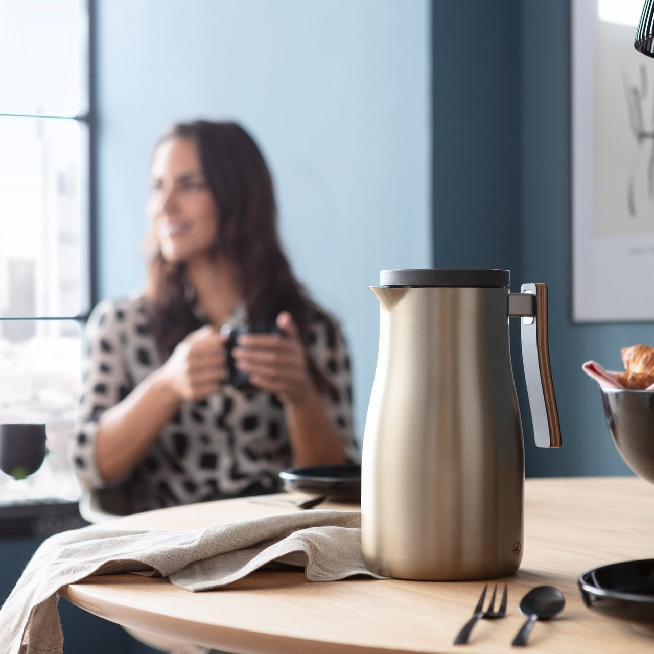 Eine Person mit langen Haaren sitzt an einem Tisch und hält einen Stift, während sie aus dem Fenster schaut. Auf dem Tisch liegen ein Notizbuch und eine Thermoskanne. Der Raum hat eine ruhige, moderne Atmosphäre mit blauen Wänden.
