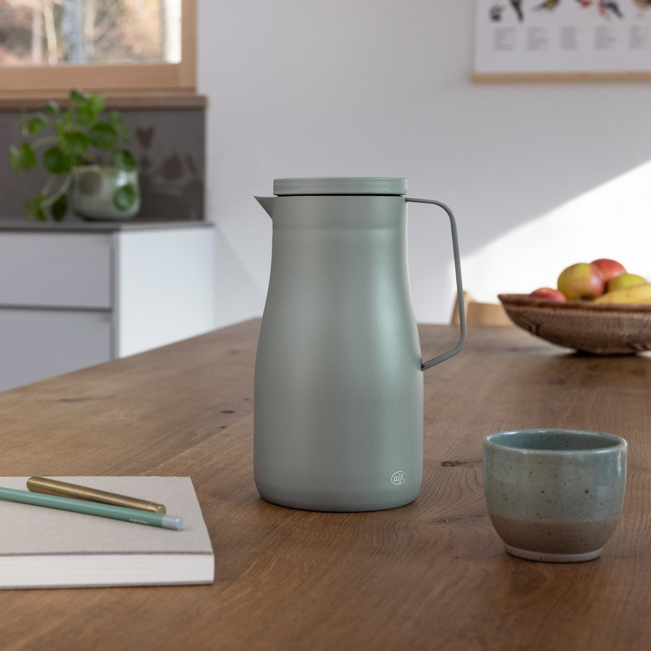 Auf dem Bild steht eine graue Thermoskanne auf einem Holztisch. Daneben befinden sich eine Tasse, ein Notizblock mit einem Kugelschreiber und im Hintergrund ein Korb mit Äpfeln. Durch ein Fenster fällt natürliches Licht in den Raum.