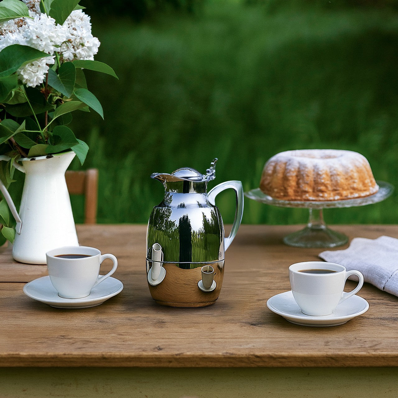 Auf einem Holztisch im Freien stehen eine glänzende Kaffeekanne, zwei Tassen Kaffee und ein Gugelhupf auf einer Glasschale. Daneben ist eine weiße Vase mit weißen Blüten. Der Hintergrund zeigt eine verschwommene grüne Landschaft.
