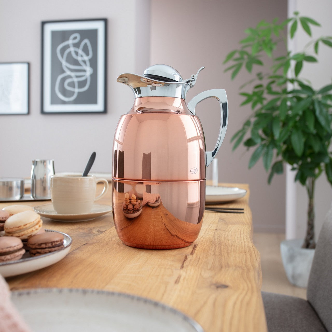 Eine glänzende, roséfarbene Thermoskanne steht auf einem Holztisch. Daneben sind eine Tasse Kaffee, Macarons auf einem Teller und eine Zimmerpflanze. An der Wand hängen zwei abstrakte Schwarz-Weiß-Bilder.