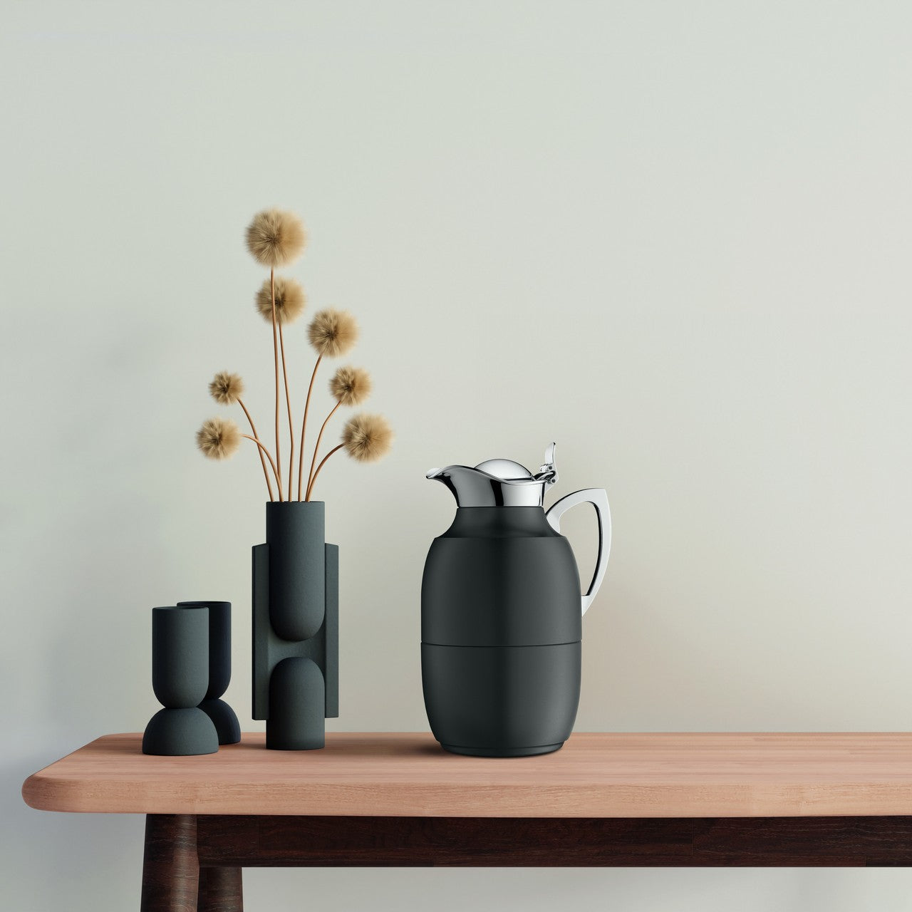 Auf dem Bild steht eine schwarze Thermoskanne mit silbernem Deckel auf einem Holztisch. Daneben befinden sich zwei modern designte, dunkelgraue Vasen mit dekorativen Trockenblumen. Der Hintergrund ist in einem hellen, einfarbigen Ton gehalten.