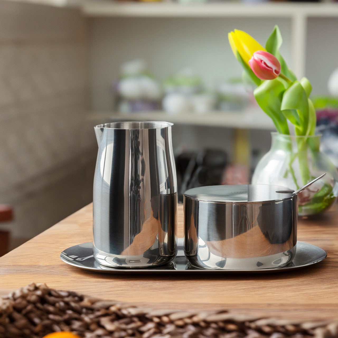 Auf einem Holztisch stehen ein glänzender Metallkrug und eine Zuckerdose mit Löffel auf einem Metalltablett. Im Hintergrund ist eine Vase mit gelben und pinken Tulpen zu sehen. Die Umgebung wirkt hell und freundlich.