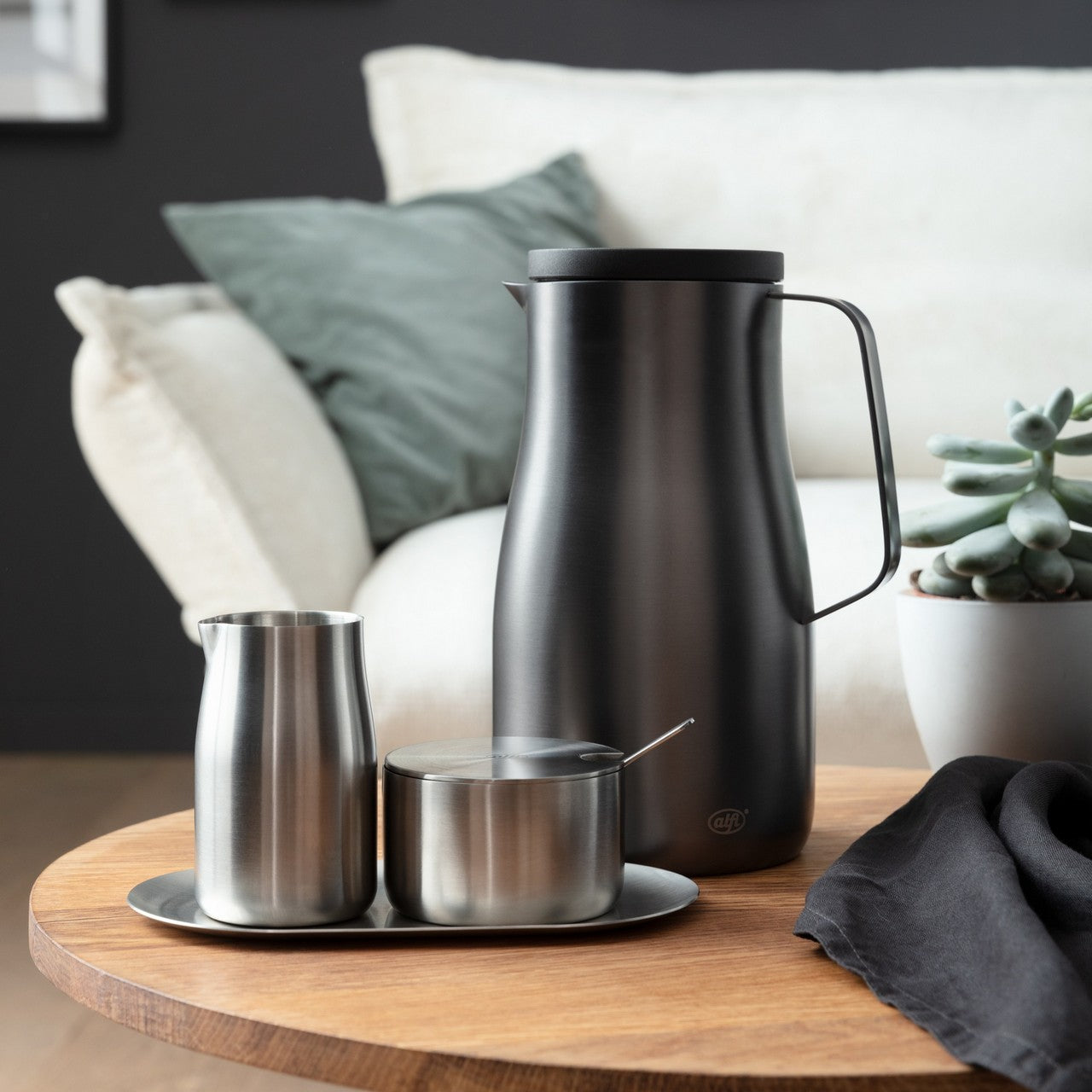 Auf einem Holztisch stehen eine schlanke schwarze Kanne, ein silberner Krug und ein Zuckerdöschen. Im Hintergrund sind ein Sofa mit grünen Kissen und eine kleine Pflanze zu sehen. Alles wirkt stilvoll und modern.
