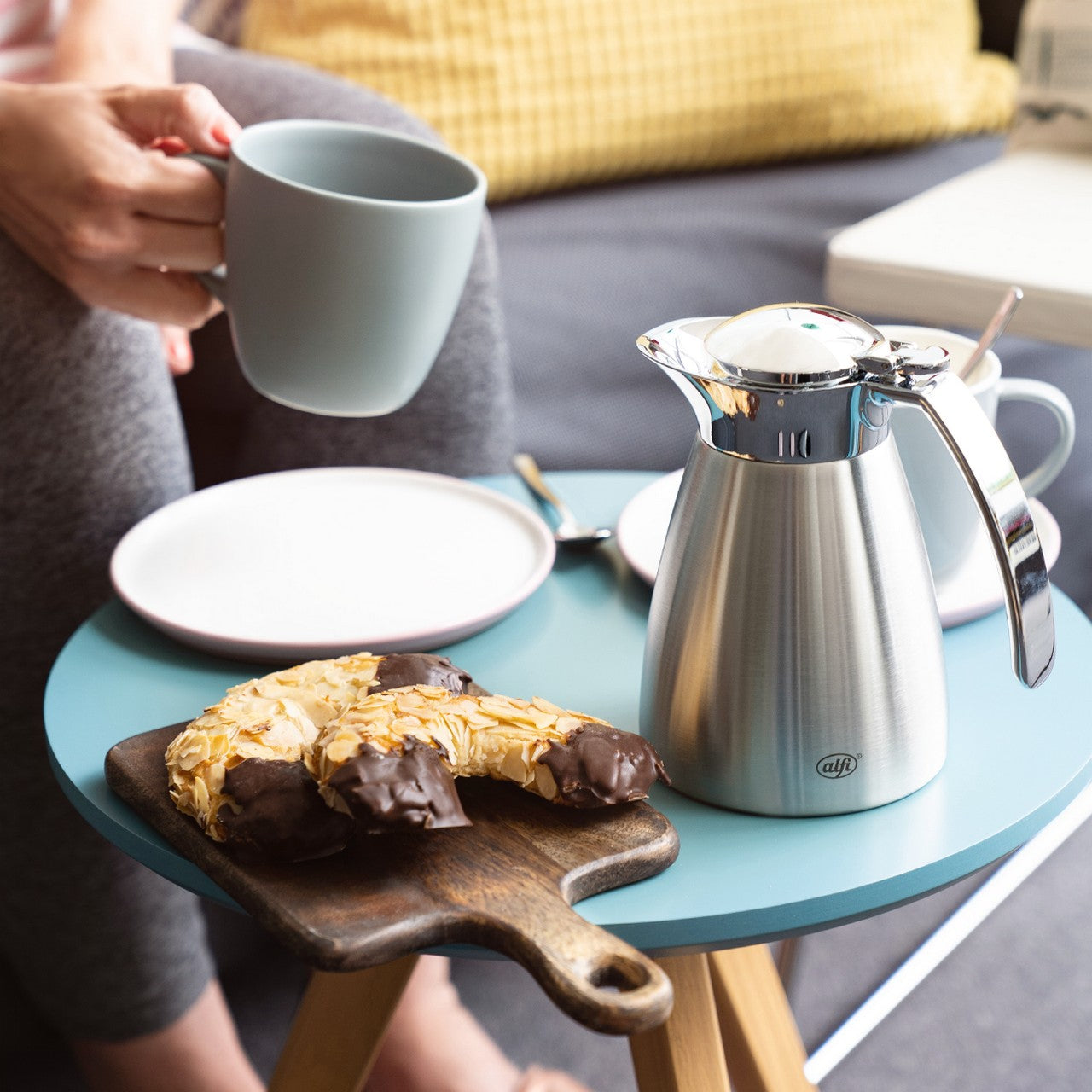 Auf einem kleinen Tisch steht eine silberne Kanne neben einem leeren weißen Teller und einem Schneidebrett mit Gebäck. Eine Person hält eine graue Tasse in der Hand. Im Hintergrund ist ein Sofa mit einem gelben Kissen zu sehen.