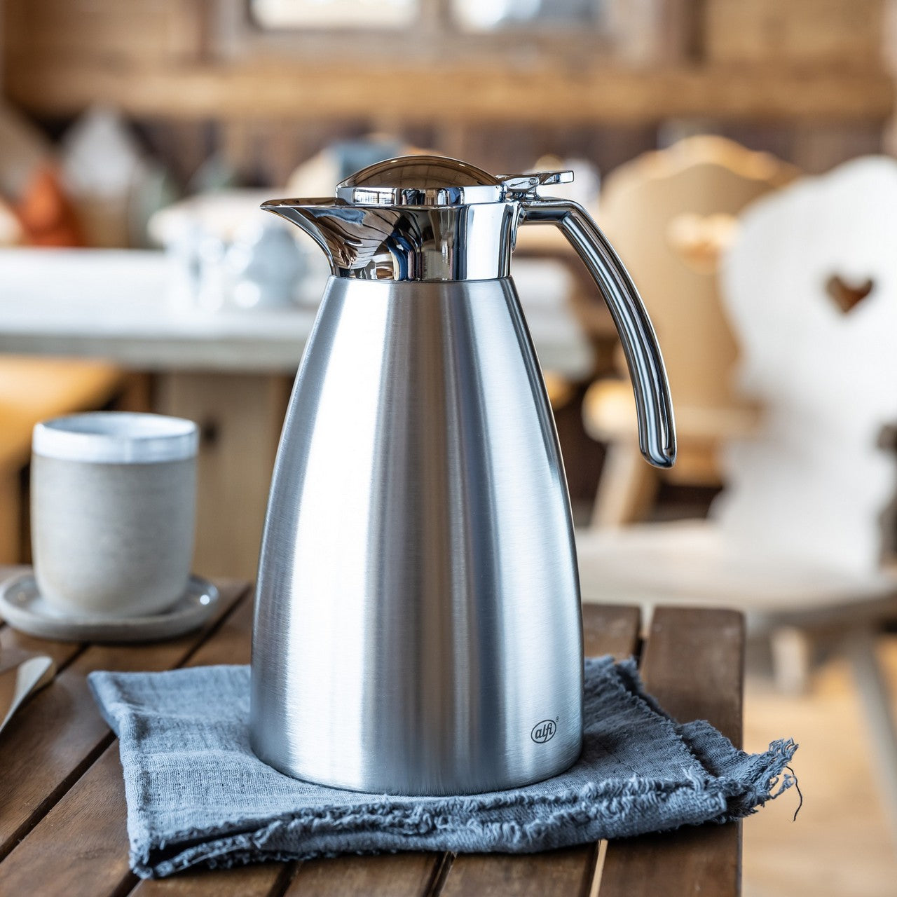 Auf dem Bild ist eine metallische Thermoskanne zu sehen, die auf einem blauen Tuch steht. Im Hintergrund befinden sich ein Kaffeebecher und Gebäck auf einem Holztisch, was eine gemütliche Frühstücksszene andeutet.
