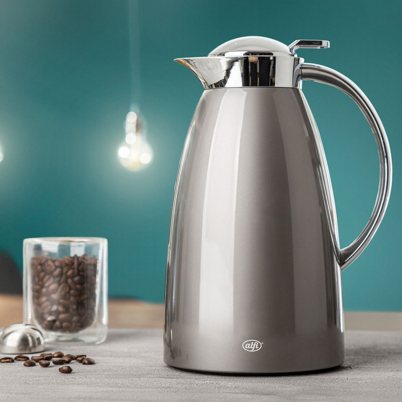 Das Bild zeigt eine silberne Thermoskanne auf einem Tisch, daneben liegt ein Teelöffel mit Kaffeebohnen und ein durchsichtiges Glasgefüll mit Kaffee. Im Hintergrund ist ein grüner Farbanstrich mit einer hängenden Glühbirne zu sehen.