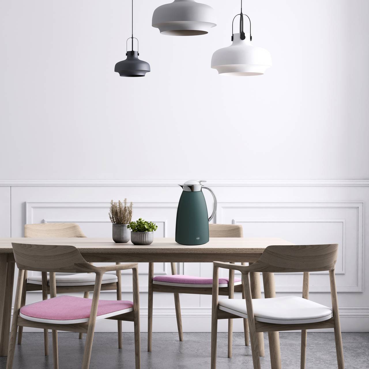 Der Raum ist minimalistisch mit einem Holztisch und vier Stühlen, zwei mit weißen und zwei mit rosa Sitzpolstern. Auf dem Tisch stehen Pflanzen und eine dunkle Thermoskanne. Drei stilvolle Lampen hängen von der Decke.