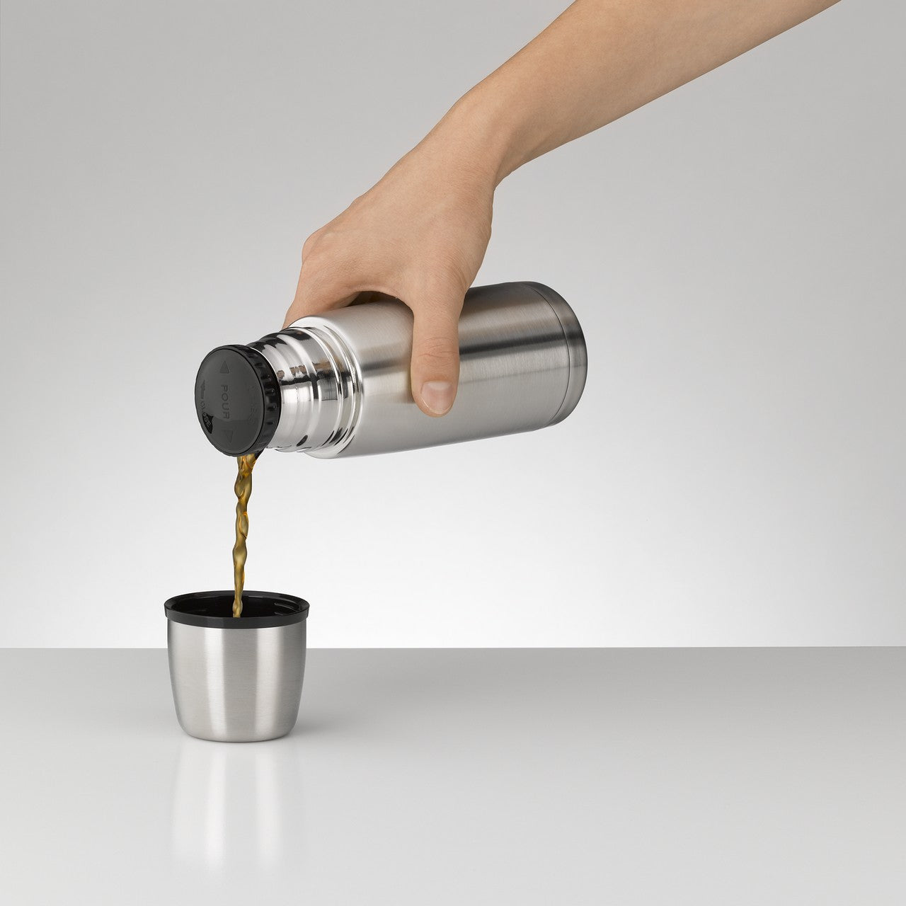 Eine Hand hält eine silberne Thermoskanne und gießt eine dunkelbraune Flüssigkeit in einen silbernen Becher. Der Hintergrund ist schlicht und grau.