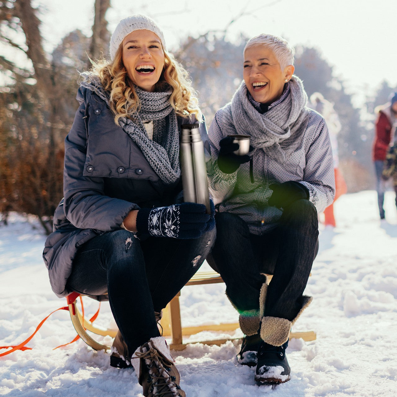 Zwei lachende Personen sitzen auf einem Schlitten im Schnee, eingepackt in warme Winterkleidung. Eine Person hält eine Thermoskanne, während die andere eine Tasse hält. Im Hintergrund sind schneebedeckte Bäume und weitere Menschen zu sehen.