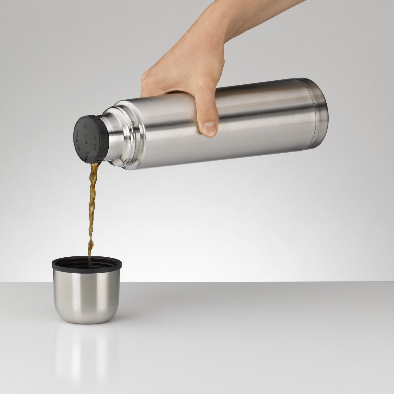 Eine Hand hält eine silberne Thermoskanne und gießt eine Flüssigkeit in einen dazugehörigen Becher. Der Hintergrund ist schlicht und grau. Die Szene wirkt minimalistisch und funktional.