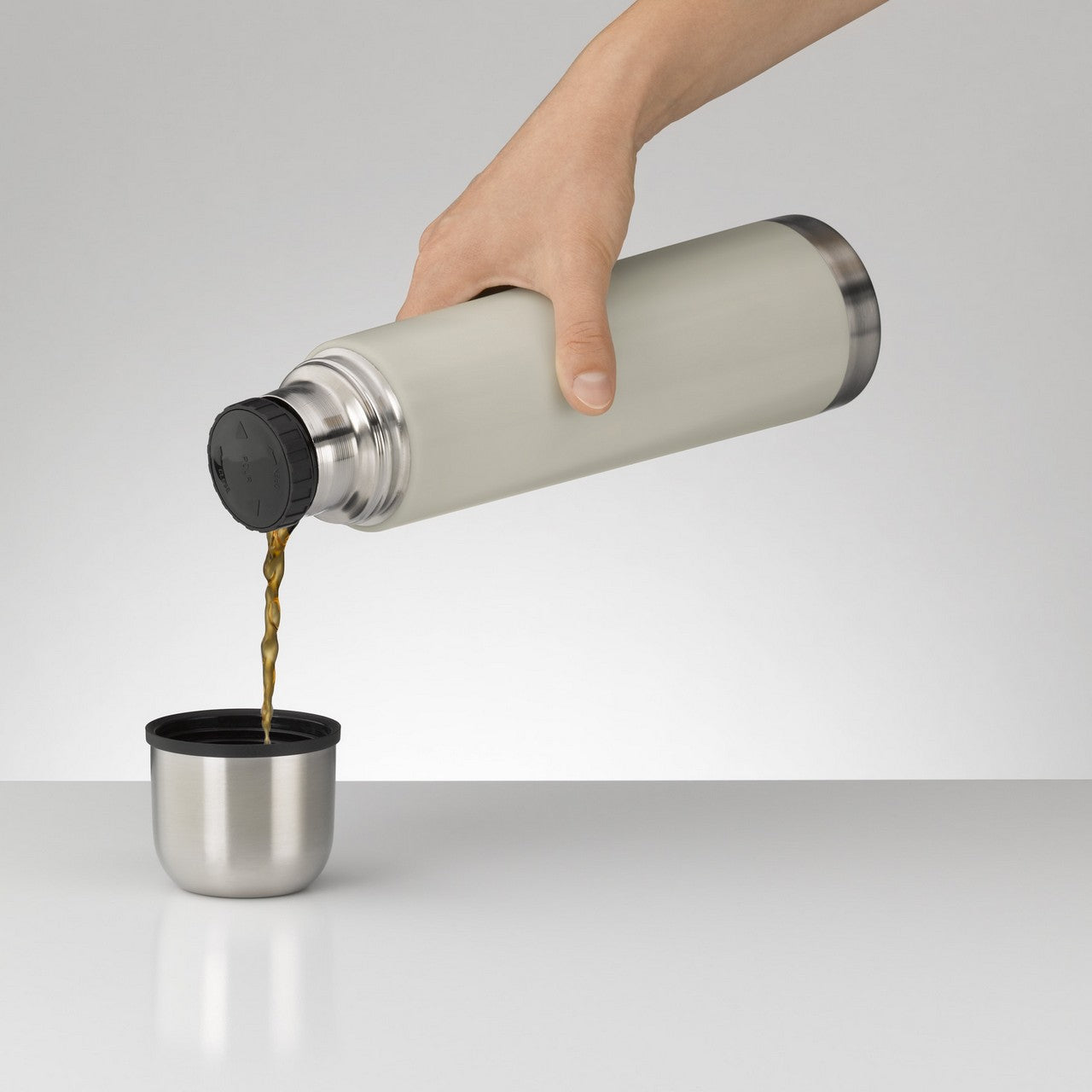 Eine Hand hält eine silberne Thermoskanne und gießt eine Flüssigkeit in einen doppelwandigen Becher. Die Szene ist vor einem schlichten grauen Hintergrund dargestellt.