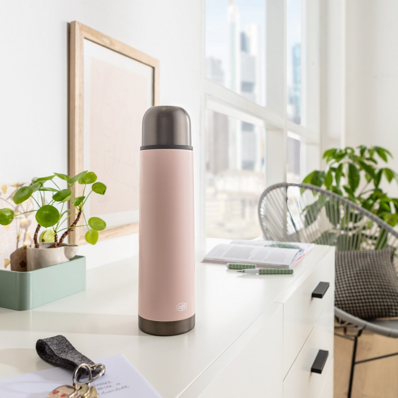 Ein moderner Schreibtisch mit rosafarbener Thermosflasche, Zimmerpflanzen und einem Computerbildschirm im Hintergrund. Alles wirkt ordentlich und hell.