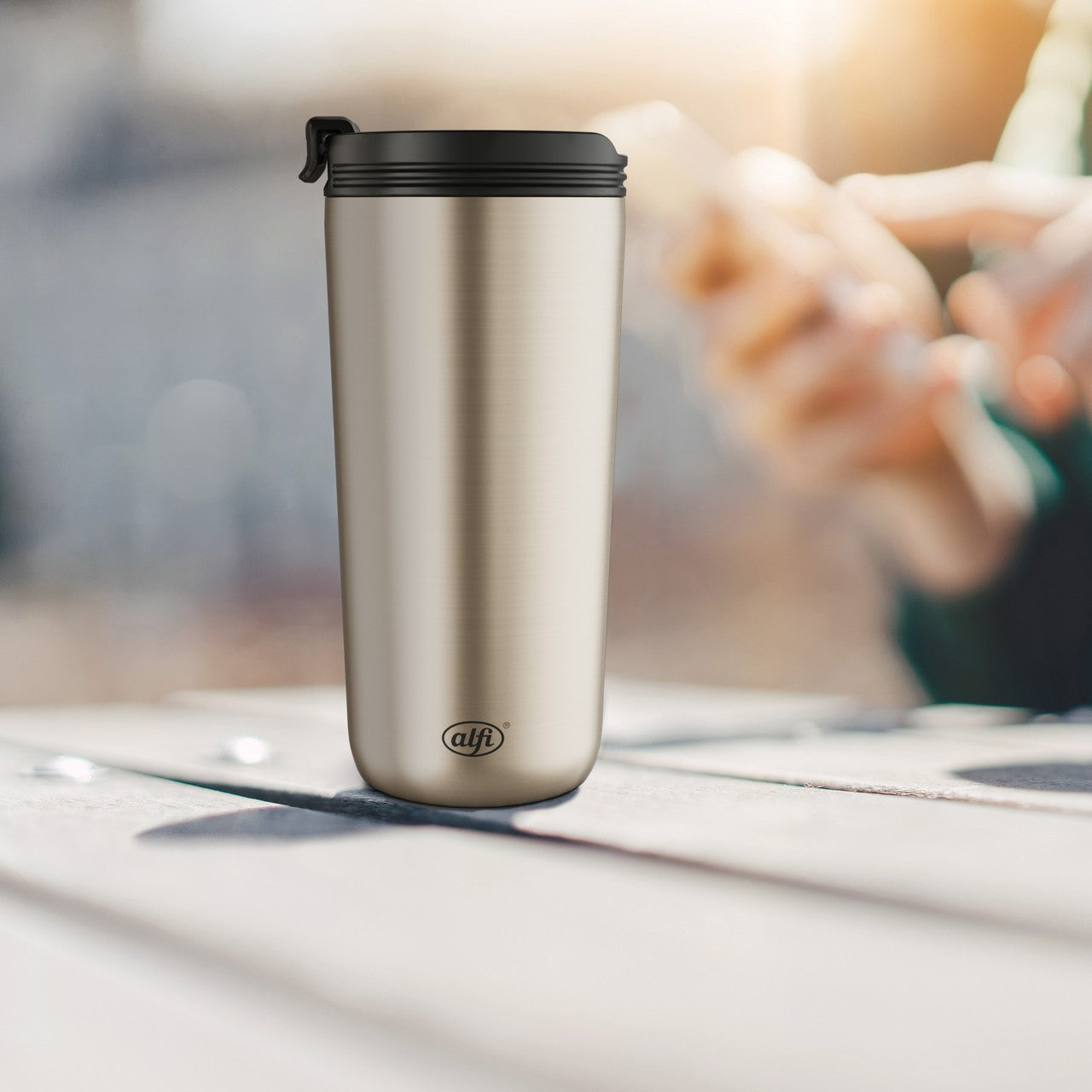 Auf dem Bild ist ein silberner Thermobecher mit schwarzem Deckel auf einem Tisch zu sehen. Im Hintergrund hält eine Person ein Smartphone in der Hand. Die Szene wirkt, als sei sie in einem Café oder einem ähnlichen Umfeld aufgenommen worden.