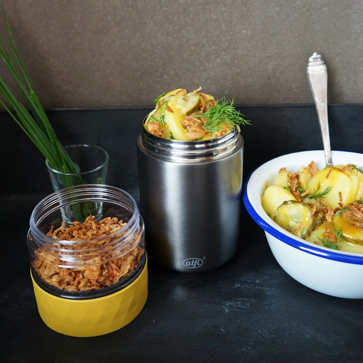 Ein Thermobehälter mit Kartoffeln und Kräutern steht neben einer Schüssel Kartoffelsalat mit Dill. Im Vordergrund ist ein Glas mit gerösteten Zwiebeln, im Hintergrund Schnittlauch.