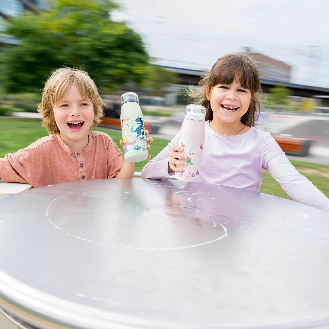 Zwei fröhliche Kinder sitzen an einem Tisch im Park und halten bunte Trinkflaschen. Sie lachen in die Kamera, während im Hintergrund Grünflächen und verschwommene Gebäudestrukturen zu sehen sind.