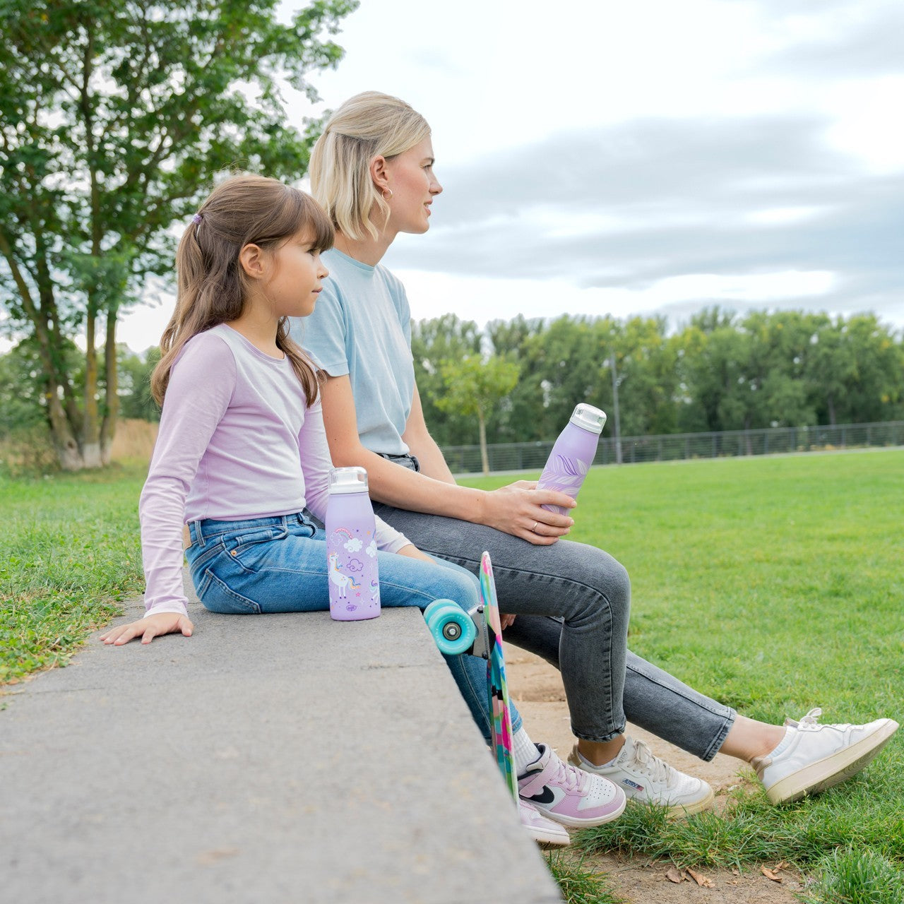 Eine Frau und ein Mädchen sitzen auf einer niedrigen Mauer im Park und halten lilafarbene Trinkflaschen. Neben ihnen steht ein buntes Skateboard. Im Hintergrund sind Bäume und gepflegter Rasen zu sehen.