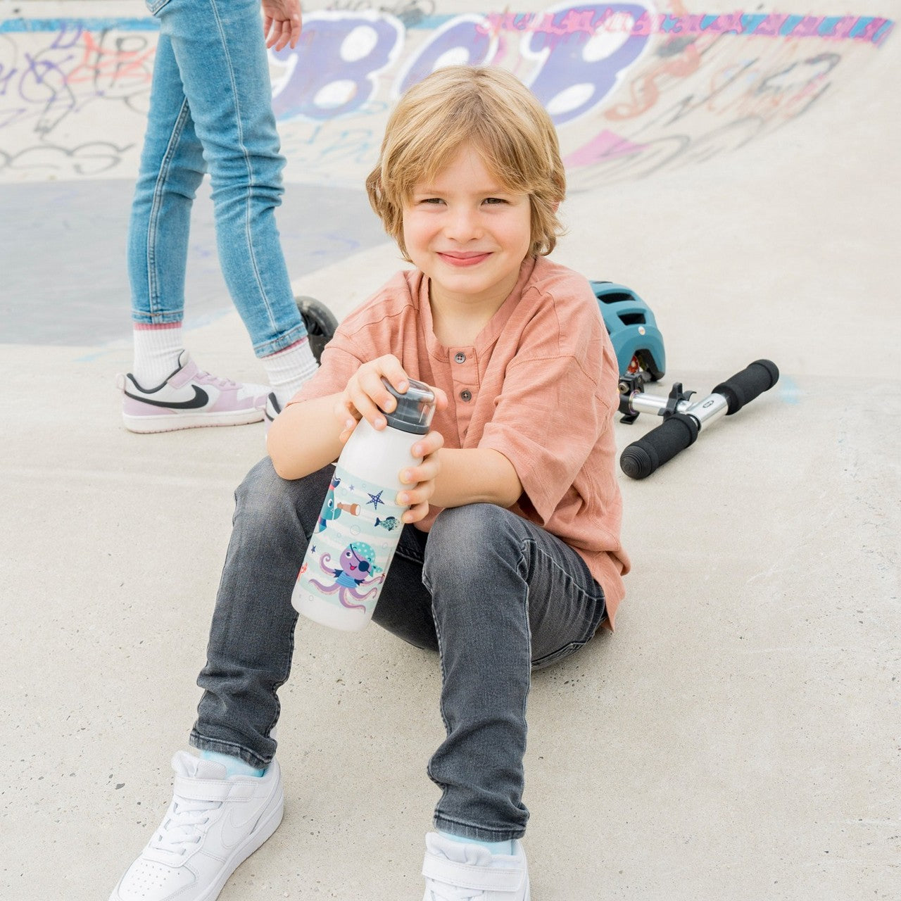 Ein Kind sitzt auf dem Boden eines Skateparks und hält eine bunte Wasserflasche, während es lächelt. Im Hintergrund sieht man einen Roller und ein Kind in Jeans sowie bunte Graffiti auf den Betonflächen.