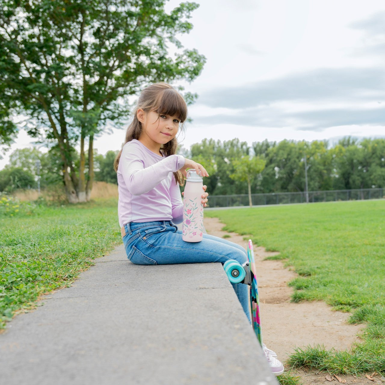 Ein Kind sitzt draußen auf einer niedrigen Mauer in einem Park und hält eine florale Trinkflasche in der Hand. Unter einem Bein steht ein farbenfrohes Skateboard. Im Hintergrund sind grüne Wiesen und Bäume zu sehen.
