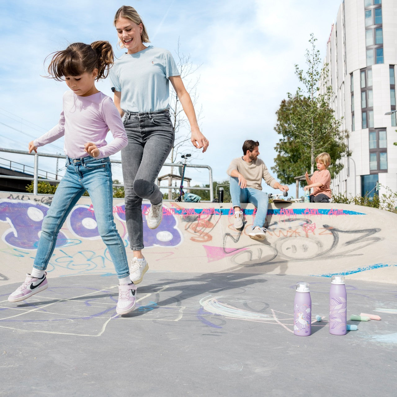 Eine Frau und ein Kind springen fröhlich auf einem Platz mit Graffiti und Kreidezeichnungen. Im Hintergrund sitzen ein Mann und ein weiteres Kind entspannt auf der Kante. Auf dem Boden stehen zwei lilafarbene Trinkflaschen.