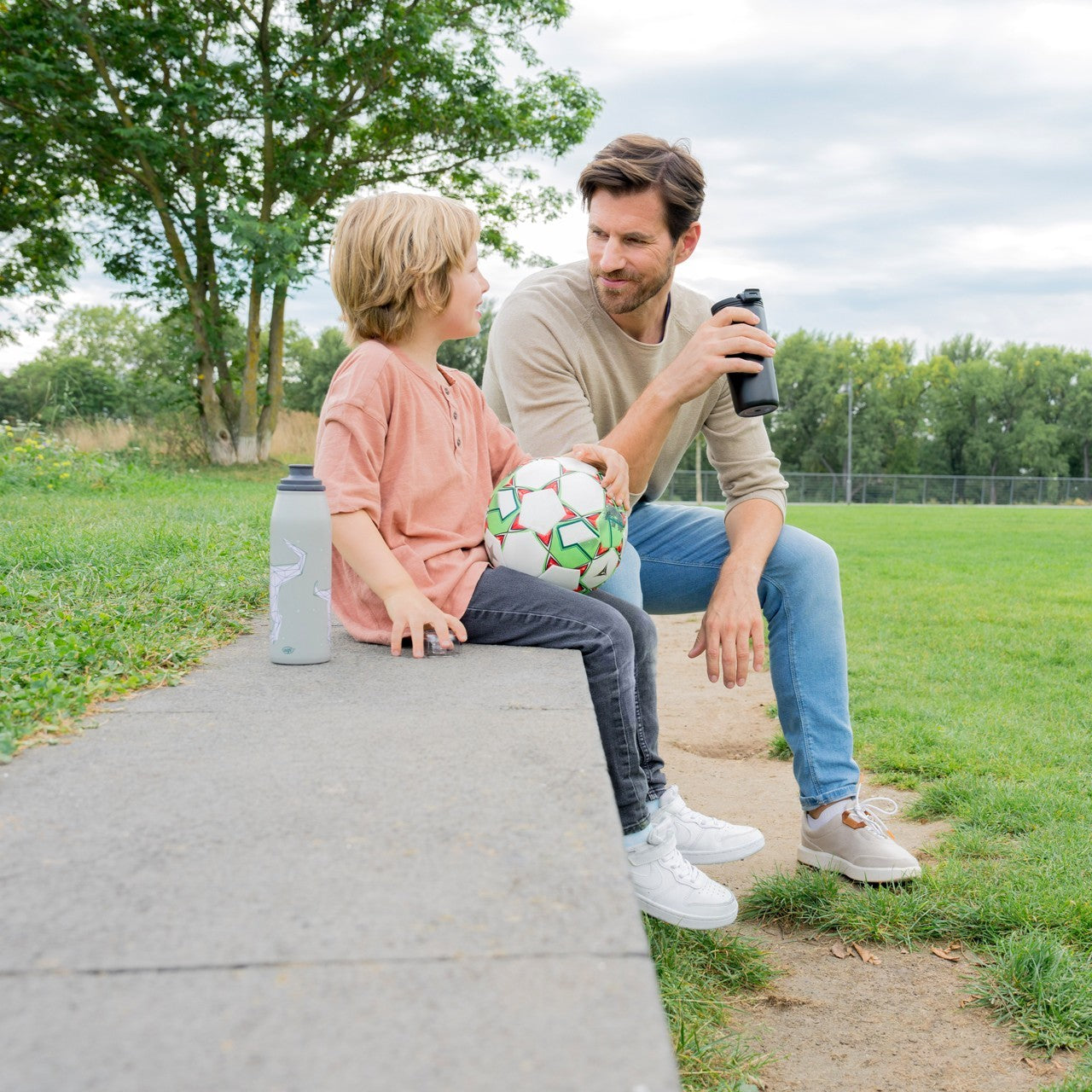 Ein Mann und ein Junge sitzen draußen auf einer niedrigen Mauer. Der Junge hält einen Fußball, der Mann hat eine Trinkflasche. Beide wirken in freundlichem Gespräch vertieft.