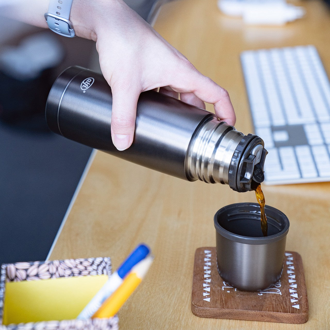 Eine Hand hält eine Metall-Thermosflasche und gießt Flüssigkeit in einen Becher, der auf einem Untersetzer auf einem Schreibtisch steht. Neben dem Becher liegen eine Tastatur und ein Stiftehalter.