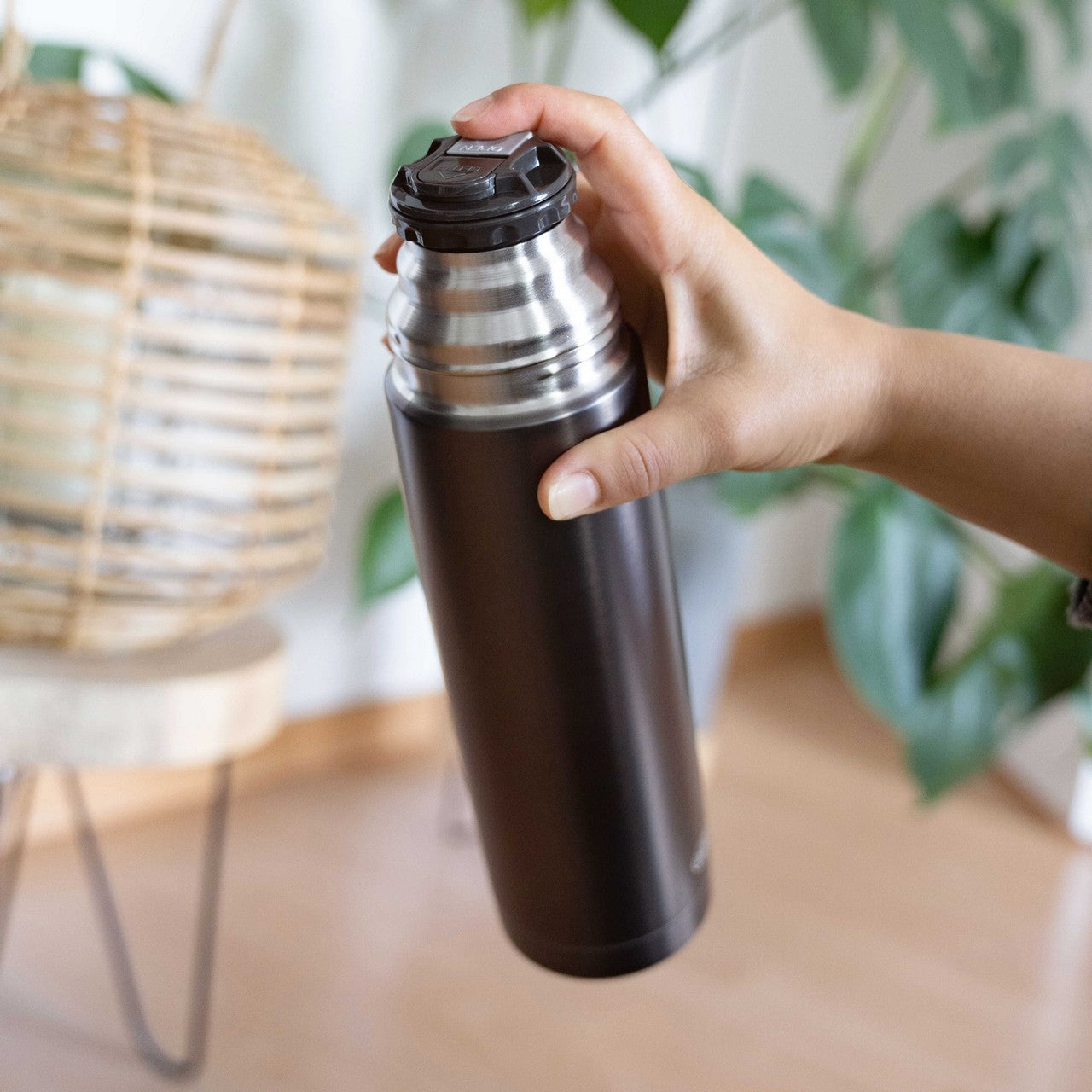 Eine Hand hält eine schlanke, dunkle Thermoskanne mit einem silbernen Deckel. Im Hintergrund sind verschwommene grüne Blätter und ein Korb zu sehen.