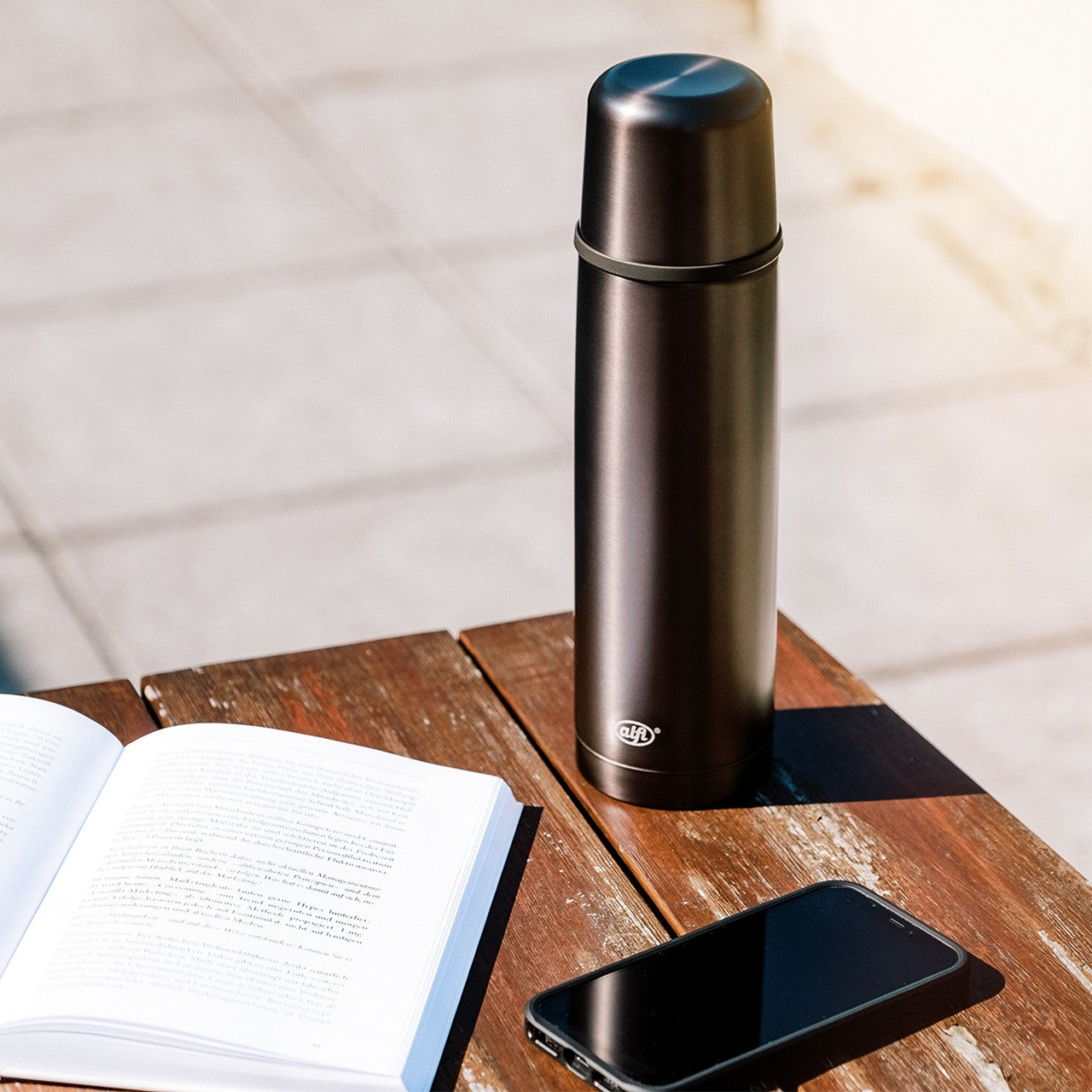 Auf einem Holztisch liegt ein geöffnetes Buch und daneben ein Smartphone. Daneben steht eine schwarze Thermoskanne. Der Tisch ist von Sonnenlicht beschienen.