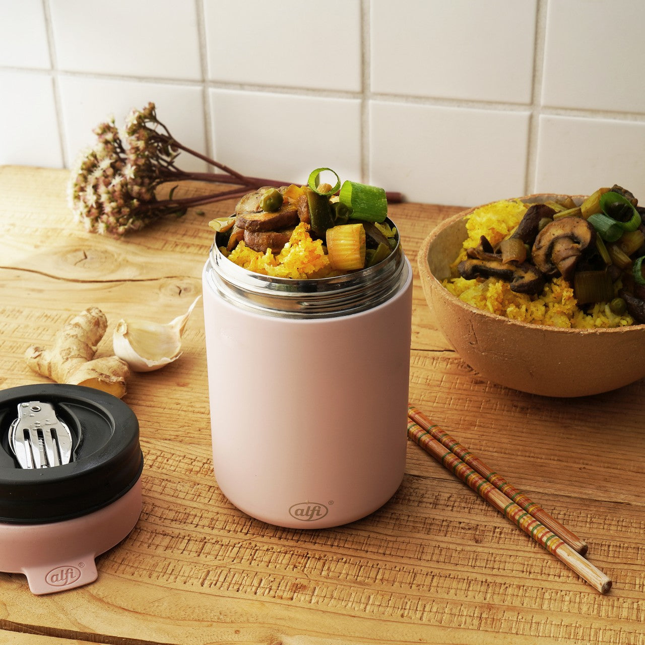 Auf einem Holztisch steht ein rosa Thermobehälter mit buntem Reis und Gemüse wie Pilzen. Daneben befinden sich eine Schale mit ähnlichem Inhalt und ein Paar Essstäbchen. Im Hintergrund sind weiße Kacheln, Knoblauch und Ingwer zu sehen.